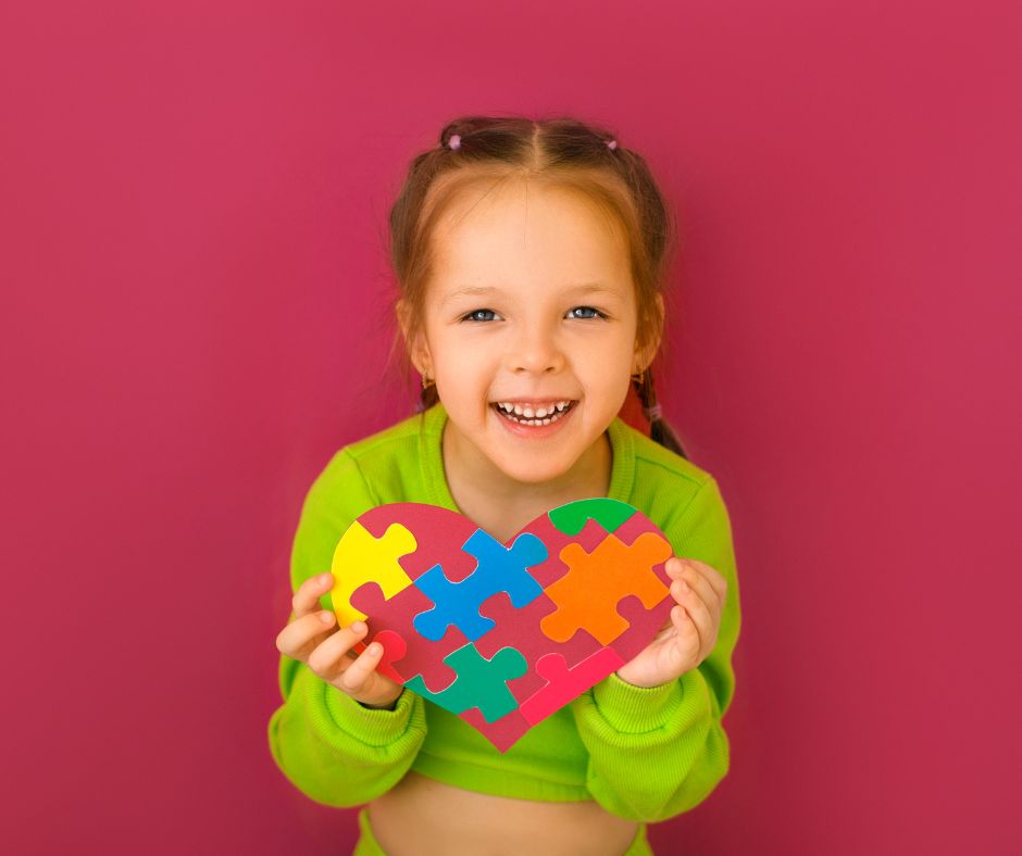 Little girl holding a heart made of puzzle pieces against a pink background.